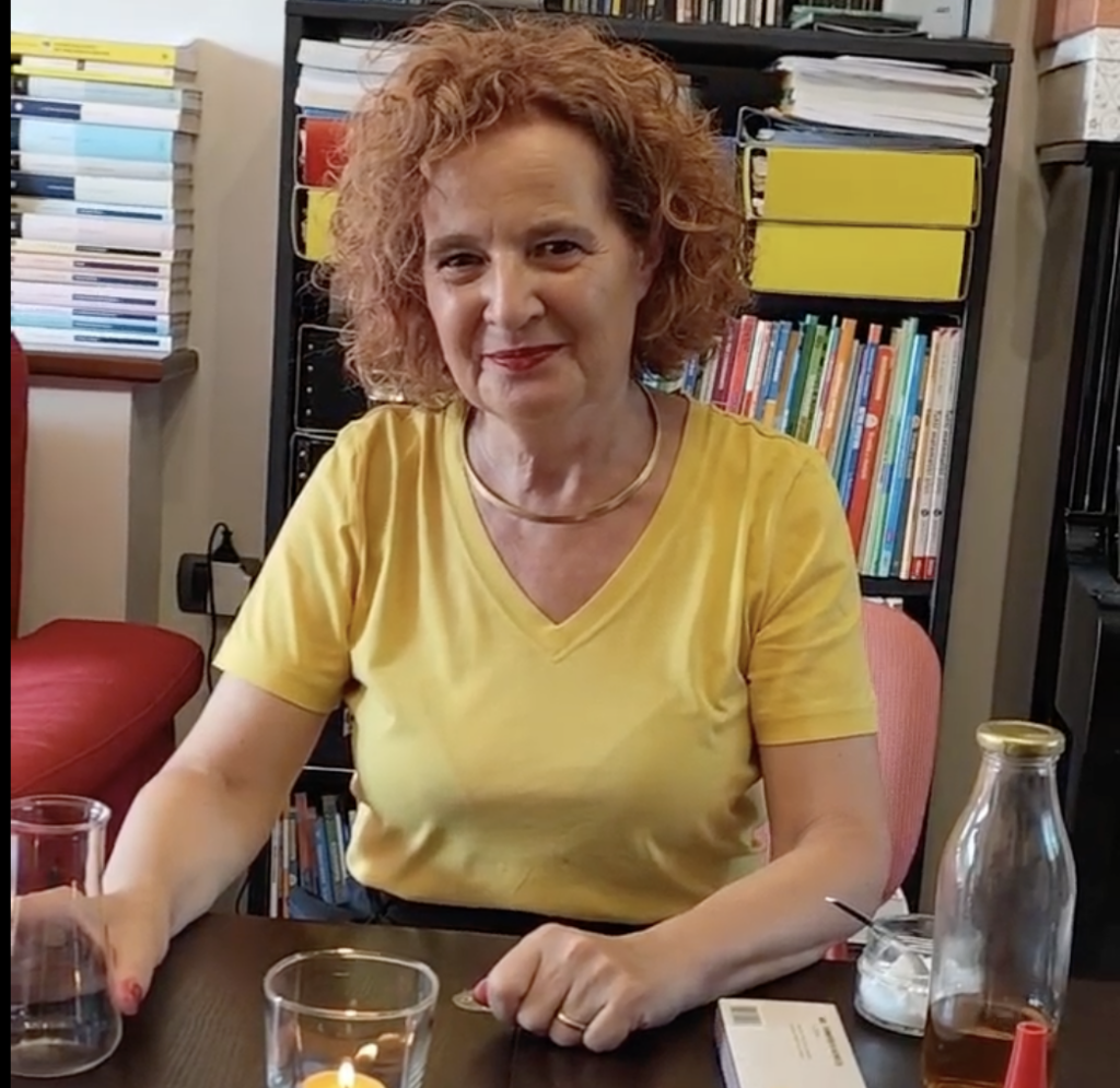 La docente Sabina Pagano seduta, indossa una maglietta gialla, ci alcune provette sul tavolo e una libreria alle spalle